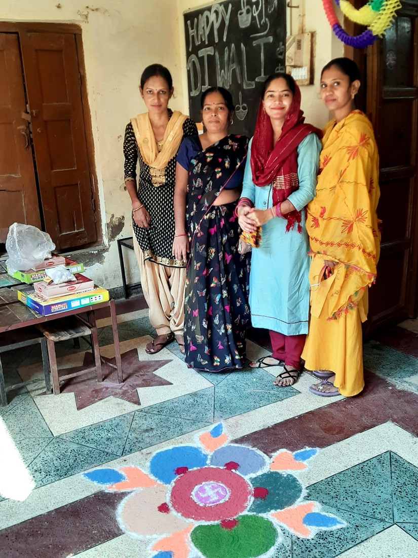 Four women teachers standing together by a Happy Diwali board and rangoli